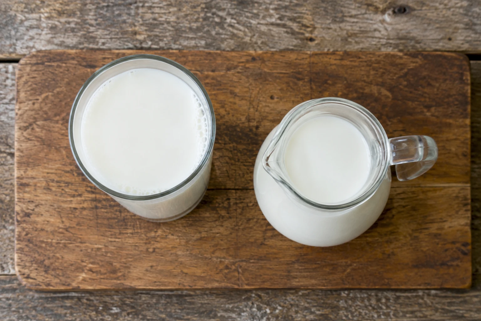 Tall glass of skim milk next to a small glass jug on a light wooden table, overhead view in soft daylight